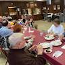 Photo of parishioners seated at a long table in the church hall. They are seated in the foreground, and the table runs from the lower right corner of the photo to the middle of the left side of the photo. The table is covered in a light red, almost maroon, plastic tablecloth, and the people are eating and socializing. The space behind the people is mostly empty except for some tables and equipment along the far wall.