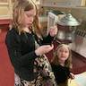 Photo of two young girls in front of the first pew in church. The older girl is on the left and standing, holding a prayer bracelet she made, while the girl to the right appears to be kneeling and is looking up at the older girl. The girl on the right is also holding a prayer bracelt.