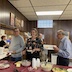 Photo from the luncheon. A man and a woman are posing while standing behind a buffet table that is covered with food atop a red, plastic tablecloth. The table is perpendicular to a wall on the left that has windows in it, through which workers in the kitchen can hand food to those attending the luncheon. Two women working in the kitchen are visible through the windows, and three men are visible behind the two people at the buffet table mentioned earlier.