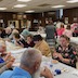 Photo from the luncheon. View of the far end of the table, which fills the lower left corner of the frame. This photo was taken from the side opposite from the previous photo. The table is (still!) covered with a white tablecloth, and people are seated on either side while eating and talking. There's a young boy in the lower right corner, facing the camera but looking just to the camera's left. A similar long table is visible in the background, and there are about six people at that table. Two men are talking at the far end, and a woman and two girls are standing with their backs facing the camera. The top of another head is partially visible between the woman and girls.