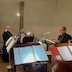 A view of the choir loft. Three women and three men are in view, standing or sitting in front of music stands as they sing.
