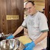 A man wearing a gray t-shirt with green letters that read Bar Harbor is standing at a table in front of a larger metal bowl. He's wearing glasses and blue food prep gloves, and there's a rectangle of dough on a slicon mat between himslef and the the bowl. He's glacing towards the camera but is not otherwise posing for the photo.