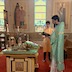 Father Aleksey stands in the right third of the frame, in front of the front of the center table, and he's facing left while holding a prayer book. An altar server stands to his right and is facing the camera. The center table contains an icon, two vigil lights, a gold cross and gold bowl, a brush, and pussy willows . The icon is surrounded by a green cloth that serves as a frame.
