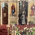 Father Aleksey is standing in the archway of the open Royal Doors with his head down as he reads from a book. He is facing the congregation, and there are flowers and a wooden cross in the lower left corner. The cross appears to be about two feet tall.