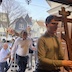 More parishioners are walking under the Epitaphios as they re-enter the church, at the end of the procession. Part of one of the wooden doors is visible in the upper right, and neighborhood houses and cars fill the background. A man along the right edge of the frame is carrying a large, wooden cross. It's the same cross that was on the center table in an earlier photo, and it appears to be a bit taller than two feet. Perhaps three?