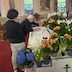 A man in the center of the frame is folding (or unfolding! I'm not sure) a white cloth while leaning over the tomb of Christ, which is surrounded by white, orange, and blue flowers, with a few dark red tulips in the mix. There's another man looking on from the bottom-left corner of the frame. Both men are wearing black shirts, and the manin the corner is wearing light beige pants. The pants of the man in the center are hidden behind the other man and are not visible.