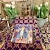 Photo of an icon of Christ on the cross. The icon is atop a small icon stand tha is covered with a purple and gold cloth with crosses on it. There are pussy willows on either side of the icon, and the icon stand is on a table with a similarly colored cloth. At the back of the icon stand, part of a gold cross is visible, and there are pussy willows on either side of the cross.There are also white tulips in small pots on the outside of the pussy willows, and gold candlestick holders on the outsides of the tulips.