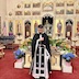 Fr. Aleksey is standing before the (not visible) congregation with his head bowed. He's wearing a black outer vestment, and a white vestment partially shown. Behind him is the Epitaphios, tomb, and flowers, with the iconostasis filling the rest of the background.