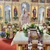 Fr. Aleksey stands in front of the open Royal Doors, facing the congregation. He's wearing white vestments with red ornamentation and is blessing the congregation using a censor in his right hand while holding a bouquet of candles in his left hand.