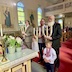 Three adults are holding large, three-branched candles while standing in front of the center table of the church. The table is located in the lower left corner of the frame, and there's a young boy holding a single, red candle in the lower right.