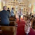 View from the back of the church as a young altar boy leads a procession down the center aisle, towards the back of the church. He's holding a large, wooden cross, and is followed, at a slight distance, by a young man carrying a gold cross on a long pole. Fr Aleksey is following behind him. Members of the congregation are seated in the pews.