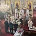 View from the choir loft, looking down towards the center table and iconostasis. Fr. Aleksey is reading from a lecturn and has altar boys with candles on either side of him. There are three men on the near side of the table holding large, three-branched candles, and there are several other men and women around the center table reading from prayer books.