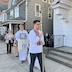 A procession around the church. An altar server is holding a large, gold cross on a long pole as he walks past the entrance to the church. Other altar servers and parishioners are seen following behind him.