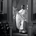 Black and white photo of Fr. Aleksey standing just inside the Royal Doors, facing the congregation and blessing them with his censer. He's facing left and fills the right half of the frame. Several vigil candles appear in the lower right corner of the photo, with the iconostas and part of a banner filling the upper left corner. Part of the iconostas is also visible along the right  edge of the frame.