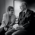 Black and white photo of a woman and a man talking while seated. She's on the left side of the frame and is gesturing with her left hand. He's holding a cup in his hands, which are resting on his lap. They're both wearing glasses.