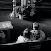 Black and white photo of a woman and a young girl — the same girl from the fourth photo — seated in a pew. The view is from behind them, and they appear in the lower right corner of the frame. There's a small table with an open pamphlet-sized book and a small stack of napkins in front of them and to their left. The top half of the photo shows two of the steps the lead towards the altar, and about a third of the way from the rights, the bottoms of a candlestand and and icon table can be seen, with a potted plant of the floor in front of and between them.