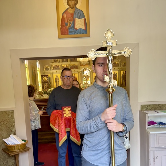 View from just outside the entrance of the church as the procession walks towards the camera, exiting the church. A man in a gray, long-sleeved shirt appears in the right half of the frame, and he's carrying a large, gold cross on a gold pole. Another man, behind him and in the center of the photo, is carrying a gold icon with a red background, and his hands are beneath a red cloth with gold trim as he holds the icon such that his hands don't come in contact with the icon. Part of a woman's face is visible as she peeks over the man's left shoulder, and another woman appears just behind the archway leading into the nave as she watches the procession moving towards her and the back of the church.