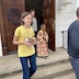View from outside the church as the procession continues to the outside. A young girl is shown center-left, and she's carrying a round loaf of holy bread that has an icon afixed to the top. To her left, in the center right of the photo, a young altar boy is wearing a reddish-purple vestment and carrying a wooden cross. There's a woman in the doorway, and she appears behind and between the two kids. Part of a man's back is visible along the right edge of the frame. Also, there's a purple wreath on the left of the double doors, and the right door is open.