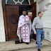 Fr. Aleksey appears in the center of the frame, in front of the entrance, as he exits the church. He's wearing white vestments with red crosses and trim, and he's holding a bouquet of candles with a red bow and some pussywillows in his left hand and a censor in his left hand. There's a man in the right half of the frame walking down the church steps.