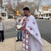 Fr. Aleksey stands at the northwest corner of the church, and he's praying while holding a large prayer book in his left hand, and he's gesturing to make the sign of the cross with his right hand. A man and woman are also visible in the photo along with some of the neighborhood houses and cars.