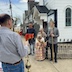 Photo of the same scene as in the previous photo except this image was taken from the perspective behind Fr. Aleksey, looking towards the church. A man's back appears in the left third of the frame, and the two men from earlier, holding the cross and icon, plus a woman holding another icon that's in a white cloth, along with the altarboy and wooden cross fill out the rest of the frame. The church is in the background behind them. A few neighborhood houses are also visible next to the church.
