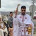 Fr. Aleksey is blessing the camera person, and a similar trail of water droplets are shown leading from bowl to brush. The woman, man, and altarboy are shown in the left half of the photo, behind Fr. Aleksey, with the neighborhood in the background and some mountains in the far distance.