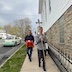 Photo of the procession along the west side of the church, with the man carrying the golden cross leading the way.  The  procession is in the center of the photo, with the church on the right side of the frame, and the street on the left. There's a pale green and yellow Volkswagon Beetle parked along the curb of the street.