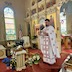 Fr. Aleksey is standing at the top of the steps in front of the open Royal Doors as he faces the congregation (not in photo). He's holding the bouquet of candles in his right hand as he reads from a small prayerbook in his left hand. The iconostas and some stained-glass windows appear in the background, and there are icon stands and flowers in the bottom of the frame.